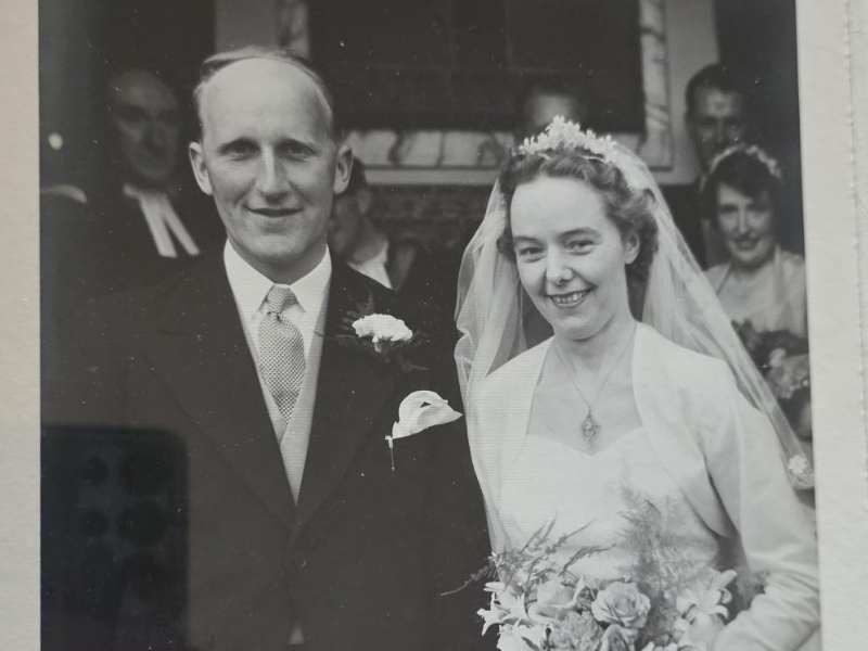black-and-white wedding photograph of a smiling bride and groom. The groom is in a dark suit with a boutonniere, and the bride wears a white gown, veil, and tiara while holding a floral bouquet. Other wedding guests are visible in the background.