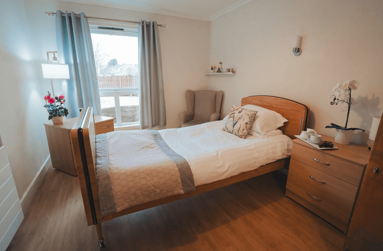 A bright, cosy bedroom featuring a wooden adjustable bed with white linens and a decorative throw. The room includes matching wooden furniture, a comfortable wingback chair in the corner, and a large window with soft blue curtains. A small tray with tea and treats sits on the bedside cabinet next to a potted orchid.