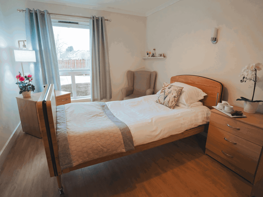 A bright, cosy bedroom featuring a wooden adjustable bed with white linens and a decorative throw. The room includes matching wooden furniture, a comfortable wingback chair in the corner, and a large window with soft blue curtains. A small tray with tea and treats sits on the bedside cabinet next to a potted orchid.