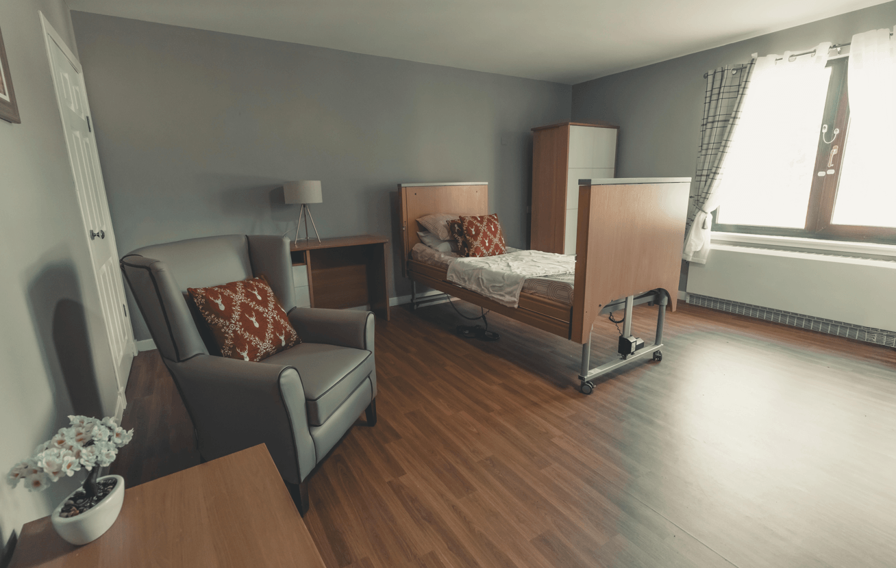 A clean, modern bedroom in a care facility. It features a high-back grey armchair with a patterned red pillow, a light wood desk with a lamp, and an adjustable hospital-style bed with a matching red pillow. The room has warm wood-effect flooring and a large window that fills the space with natural light