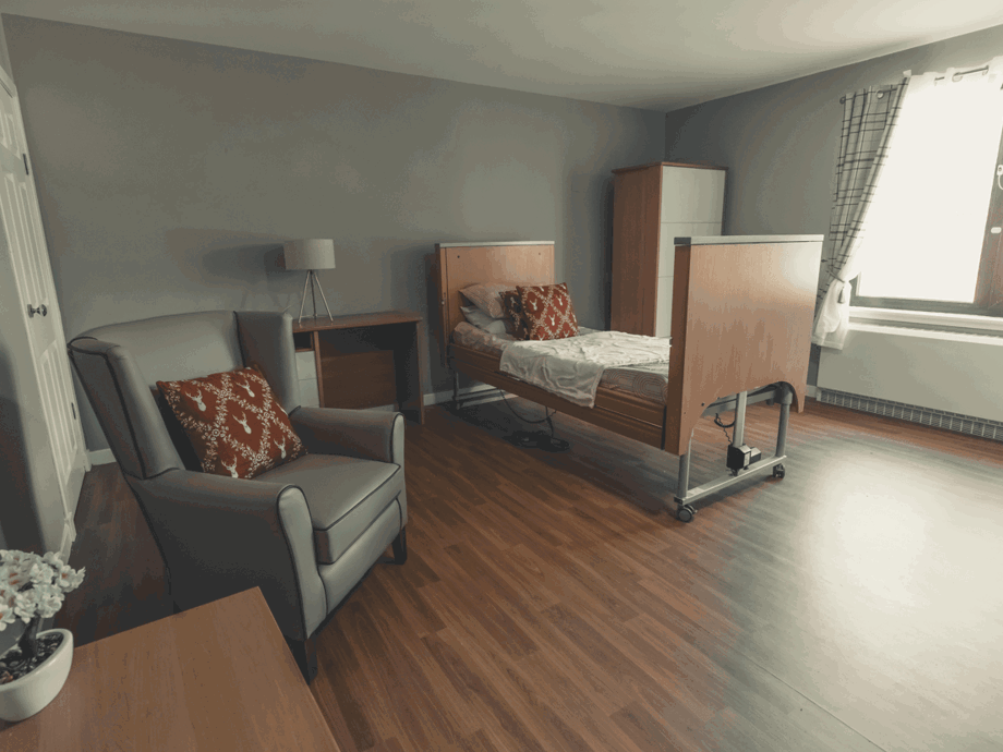 A clean, modern bedroom in a care facility. It features a high-back grey armchair with a patterned red pillow, a light wood desk with a lamp, and an adjustable hospital-style bed with a matching red pillow. The room has warm wood-effect flooring and a large window that fills the space with natural light