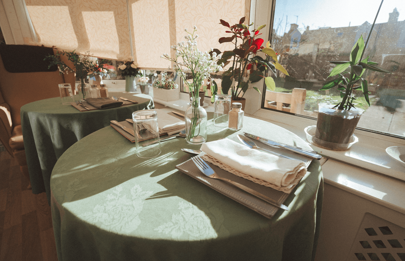 A close-up of a small dining table in a sunlit room. The table is covered with a green cloth and set with white linens, silverware, and a small glass vase of white flowers.