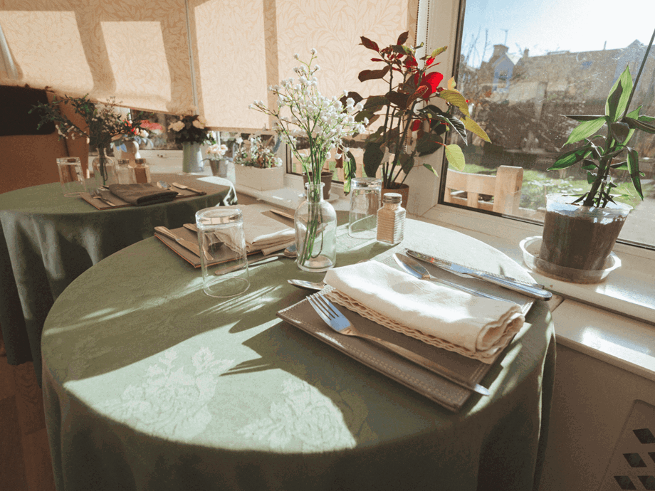 A close-up of a small dining table in a sunlit room. The table is covered with a green cloth and set with white linens, silverware, and a small glass vase of white flowers.