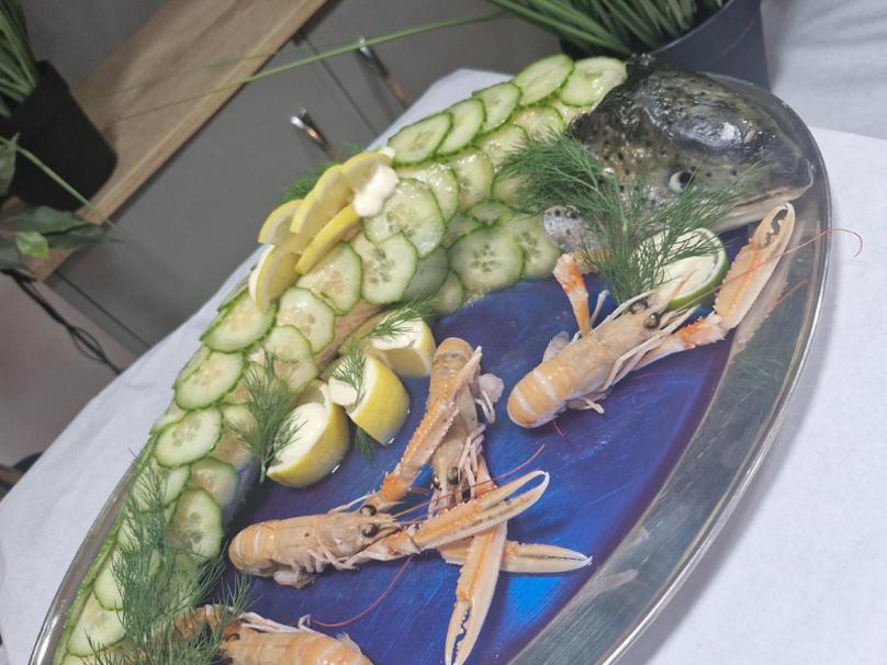 A close-up of a large silver platter featuring fresh seafood, including whole prawns (langoustines) arranged on a bed of ice. The platter is garnished with slices of cucumber, lemon wedges, and dill