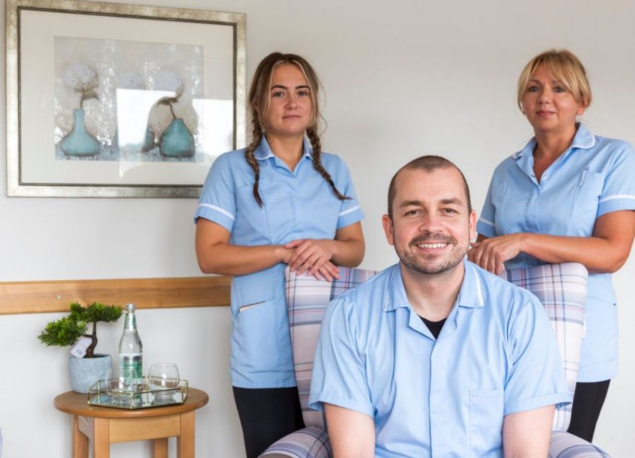 Two staff members stand behind a third staff member who is sitting in an armchair within the homes communal lounge. The environment is modern and bright