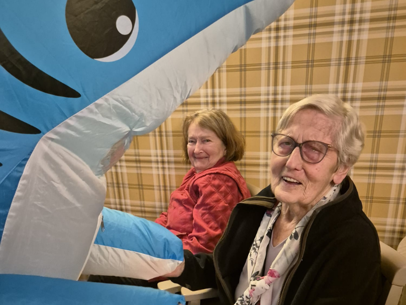 Two older women are seated next to each other, smiling warmly. A large blue and white inflatable shark is partially obscuring the foreground - dancing as part of a themed activity.