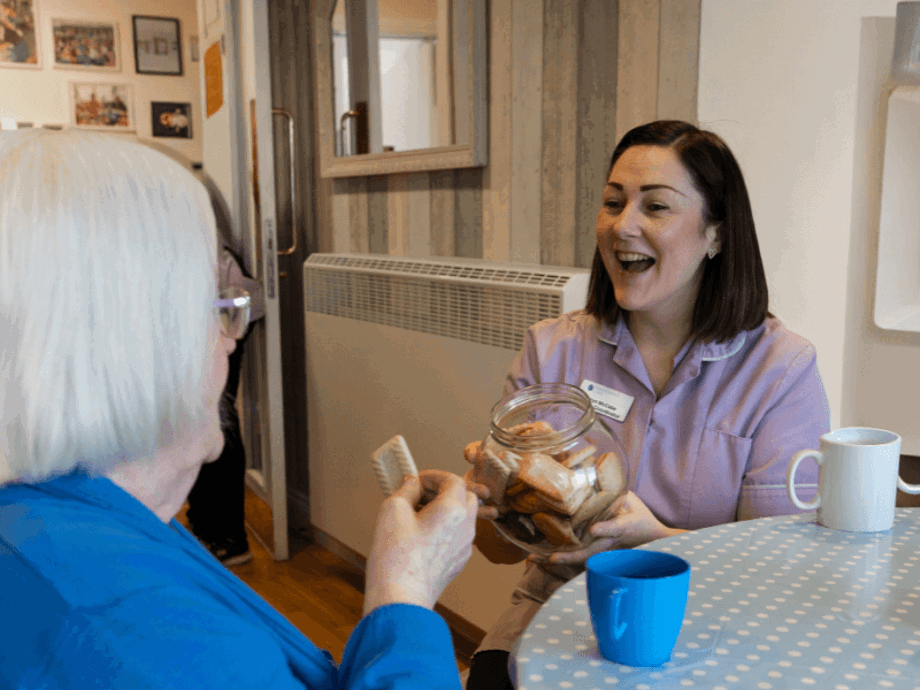 A female caregiver in a purple uniform laughs brightly while holding out a large glass jar of biscuits to an elderly woman seated at a table. A blue mug and a white mug sit on the polka-dot tablecloth between them.