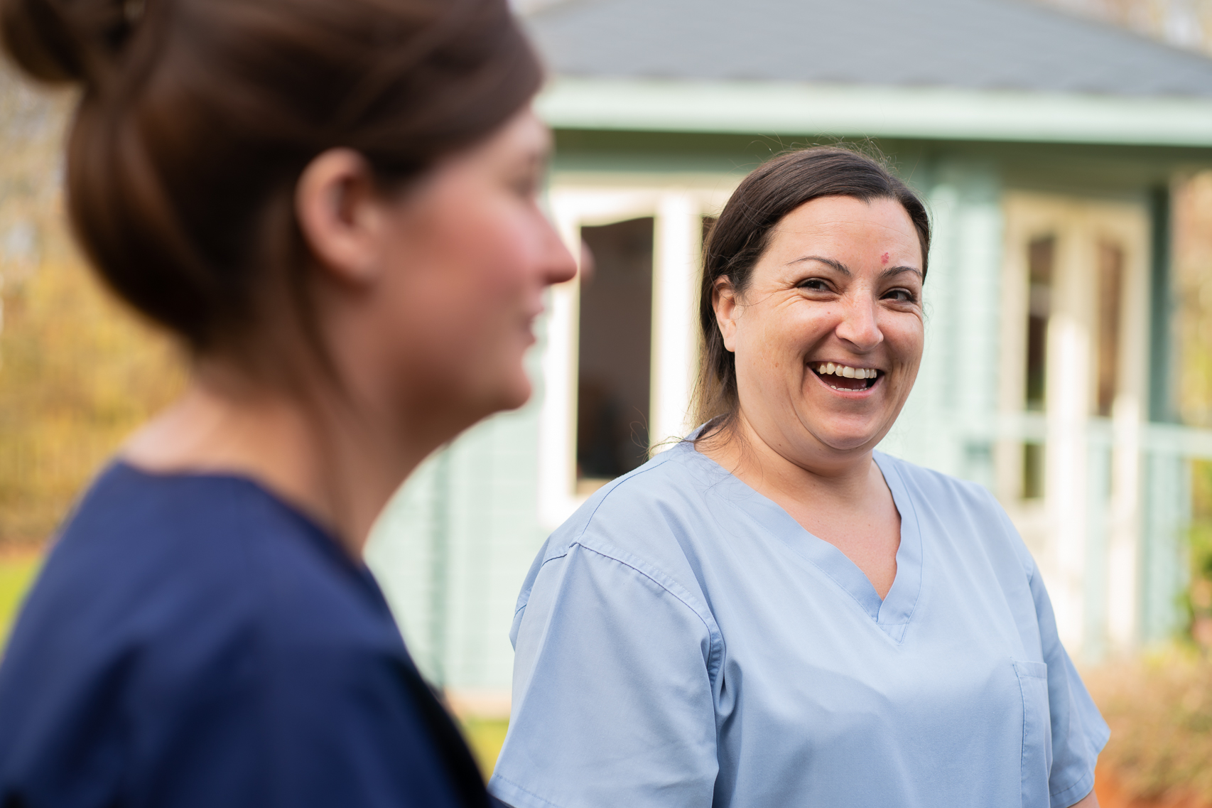 Two staff members stand in the garden. Only one is in focus, she is looking over and laughing