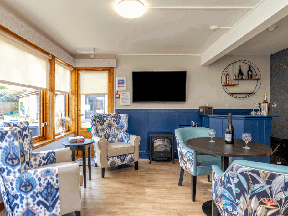 A cosy lounge and bar area with a blue wood-paneled wall, a wall-mounted TV, and an electric stove fireplace. Two wingback chairs with blue and white patterns sit near a window, while a small bar and a round table with blue floral chairs are positioned on the right.