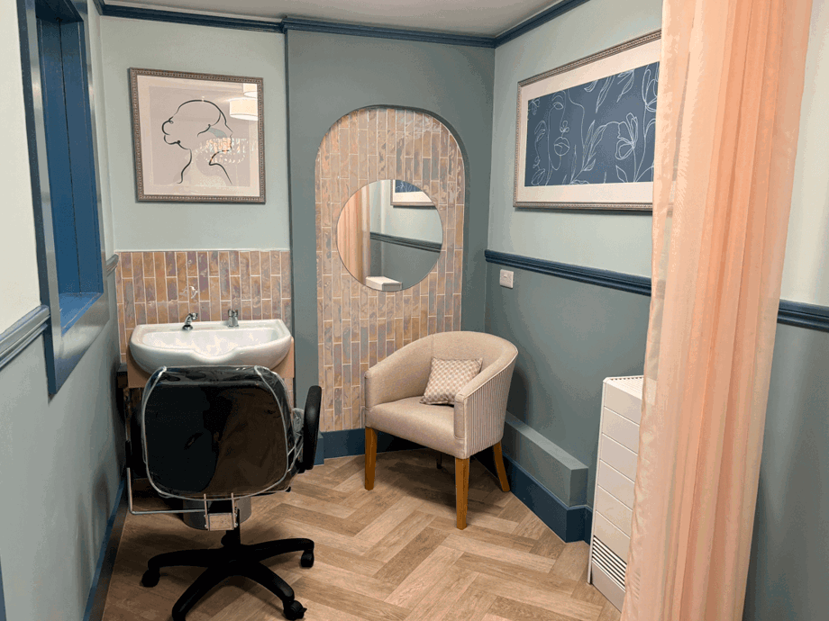 A light blue-painted treatment room with a tiled accent wall around an arched alcove and mirror. The room has a sink, a black adjustable chair, and a small cream-coloured armchair.
