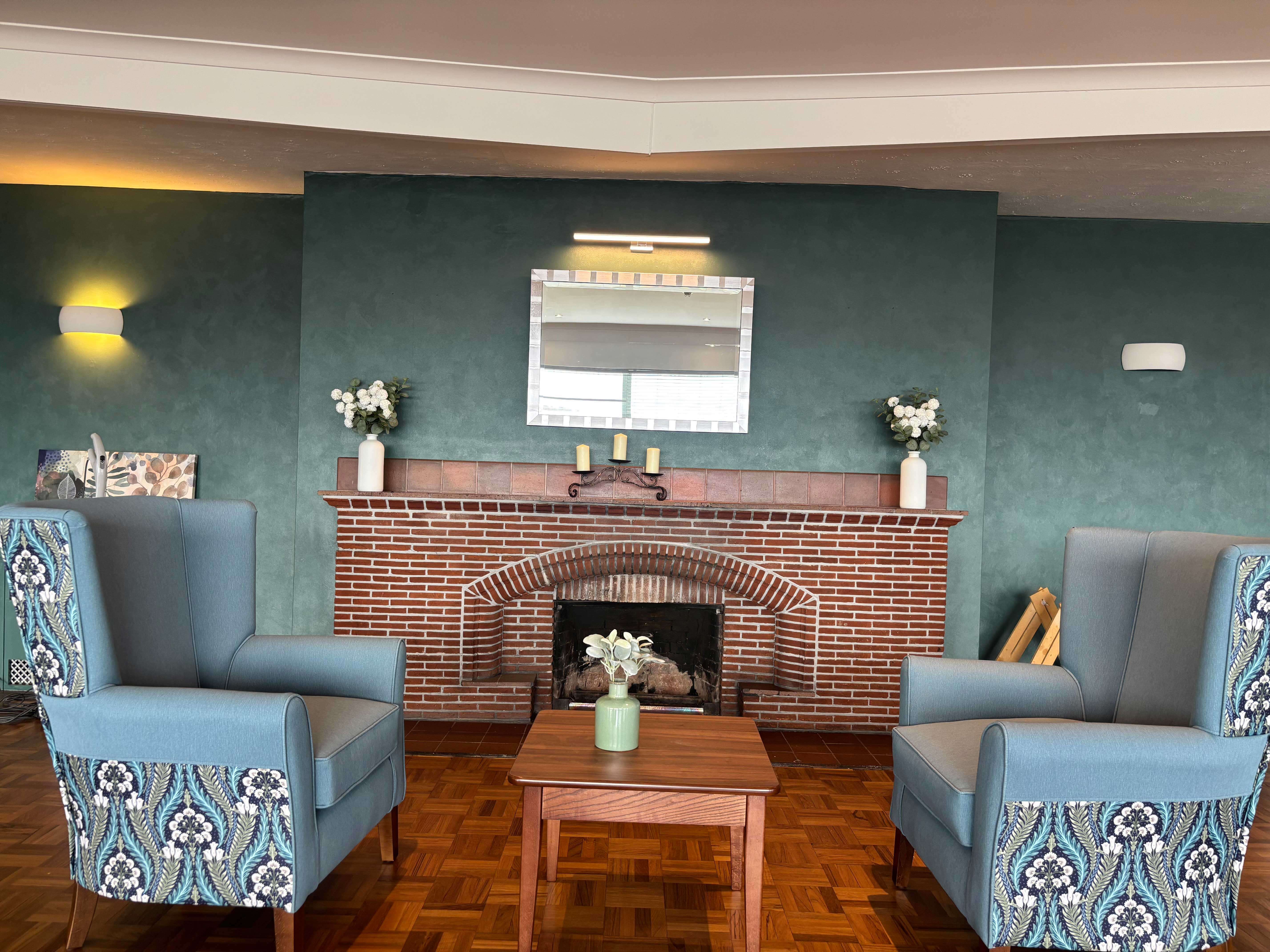 A cosy sitting area featuring two large, high-backed armchairs upholstered in solid light blue with a vibrant blue-and-white patterned base. The chairs face a small wooden coffee table and a brick fireplace set against a dark teal accent wall.