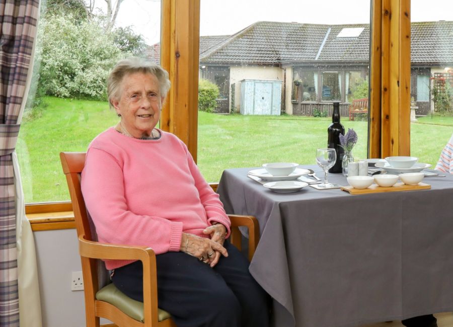 A care home resident sits in a wooden chair at a table in a care home dining room. A large window behind her offers a view of the garden.
