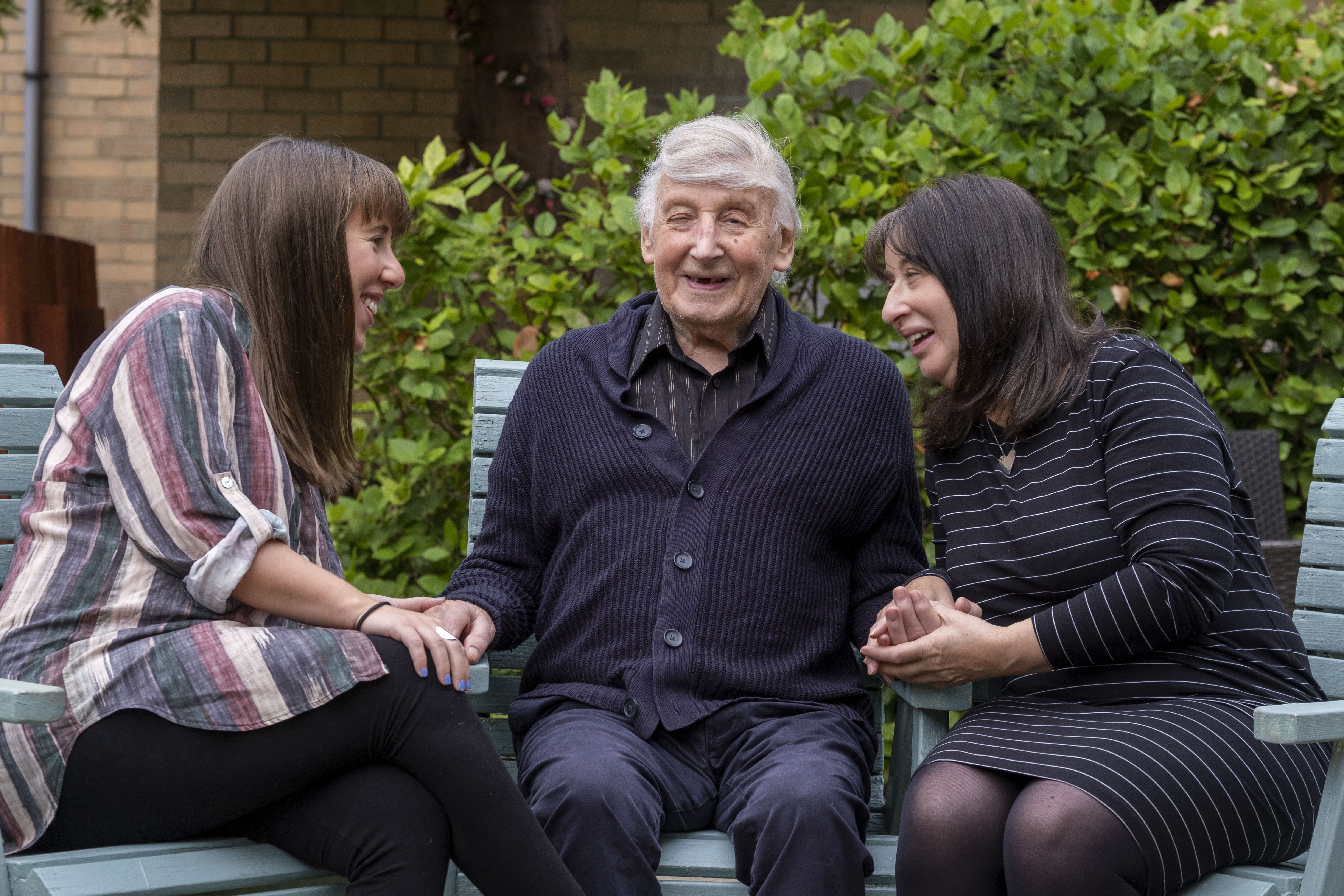 3 generations of a family are sitting in the garden on blue deck chairs as the family visit their father and grandfather respectively 