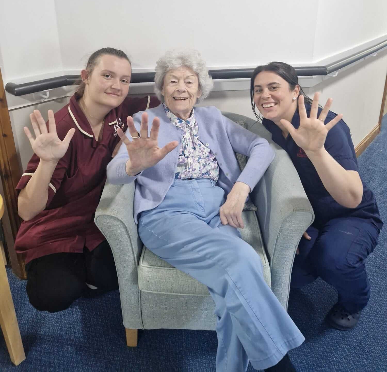 two staff members crouch next to a resident sat in a bucket chair. The three people raise up an extended hand each - showing 5 fingers for their recent 5 star report from the care inspectorate