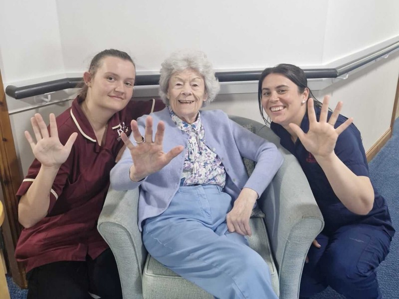 two staff members crouch next to a resident sat in a bucket chair. The three people raise up an extended hand each - showing 5 fingers for their recent 5 star report from the care inspectorate