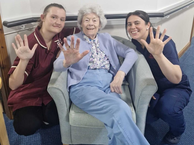two staff members crouch next to a resident sat in a bucket chair. The three people raise up an extended hand each - showing 5 fingers for their recent 5 star report from the care inspectorate