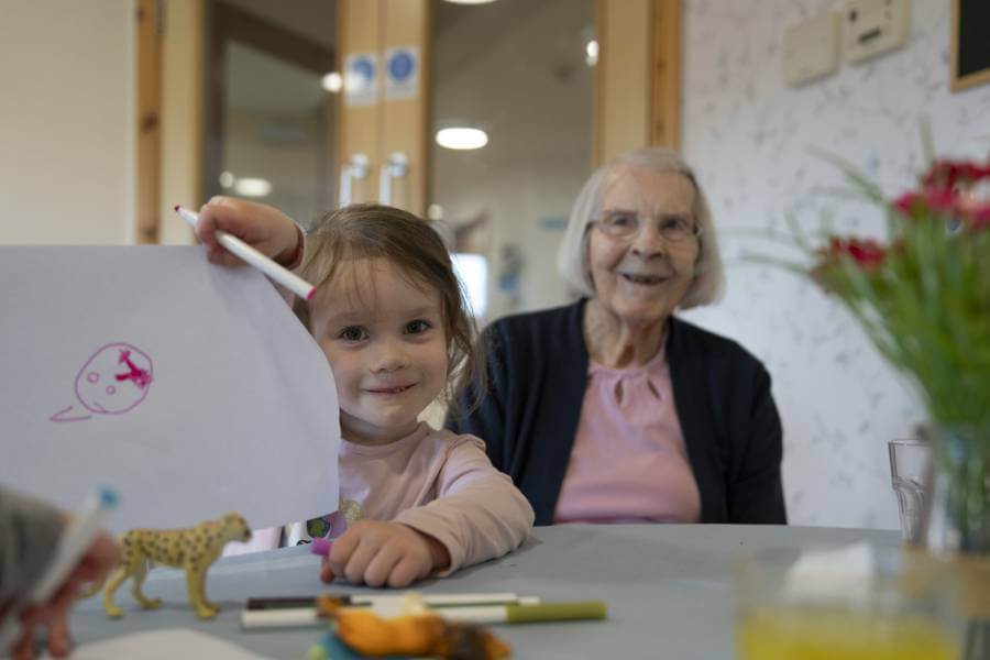 A nursery child holds up her latest drawing. A care home resident sits behind offering an encouraging look