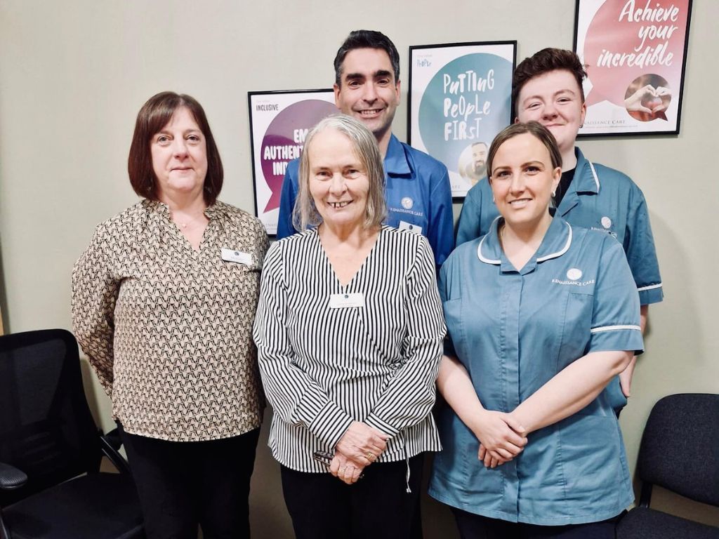 A group of five adults, three women and two men, are standing and smiling in an indoor care home setting, with values posters on the wall behind them.
