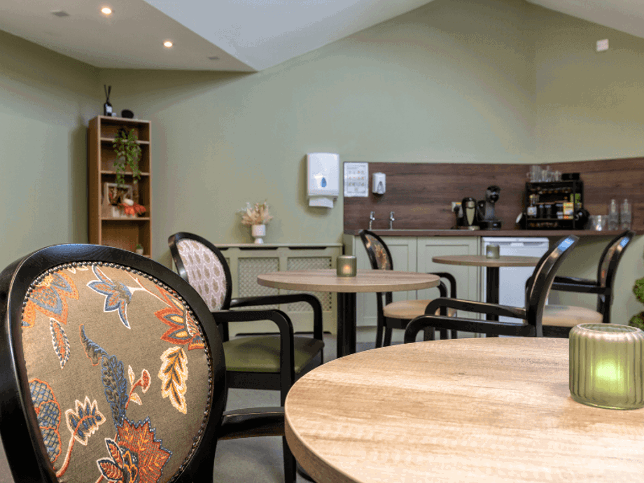 A quiet café corner featuring small round wooden tables and black-framed armchairs with floral patterned backs. In the background, there is a kitchenette area with a coffee machine, a small refrigerator, and a shelf with plants. The walls are a soft sage green.