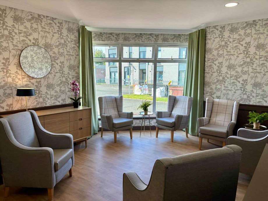 A resident lounge comprised of a sideboard and a selection of wing-back chairs. The room is calming with neutral and green colours and large bright windows.
