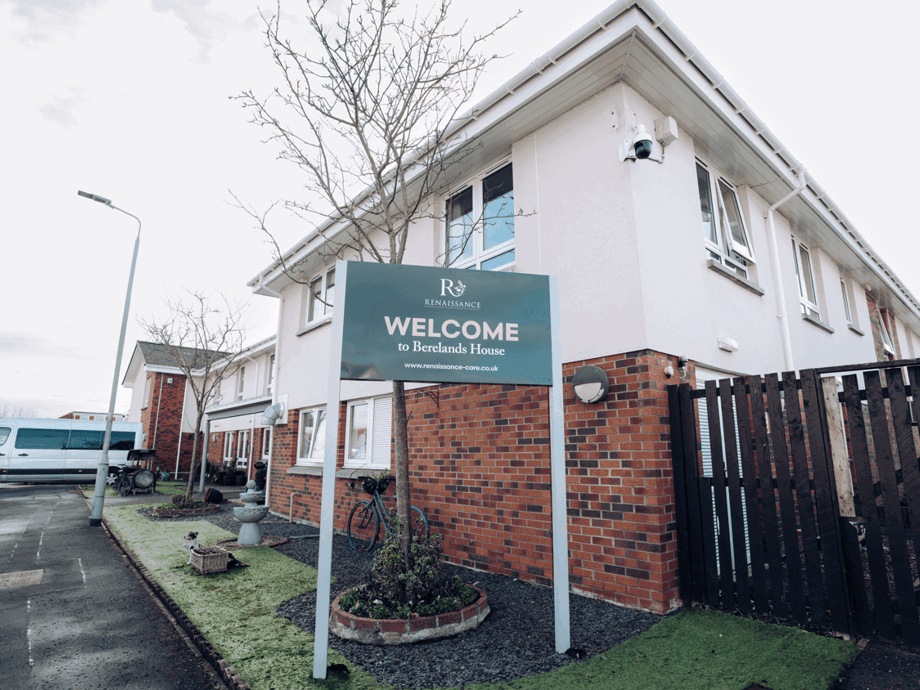 A two-story modern purpose-built building with white siding and brick accents. A green sign in the foreground reads "WELCOME to Berelands House" under the Renaissance Care logo. A small garden area with a fountain and a bicycle is visible in front of the building.