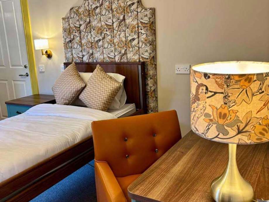 Image of a modern bed with white bedding, a wooden side table with a lit lamp, and visible floral patterns in the room