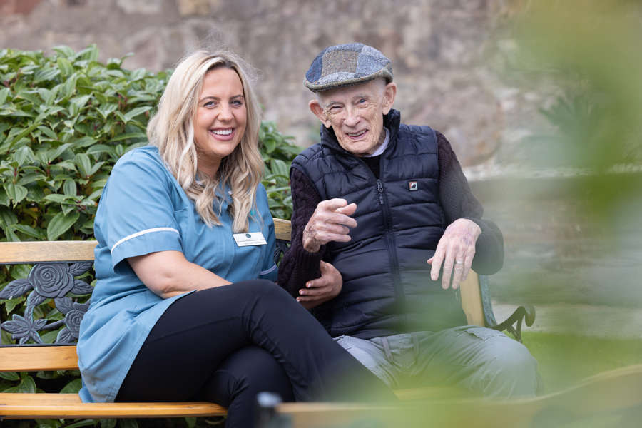 A staff member and a resident sit beside each other on a bench in the care homes garden. They are laughing together