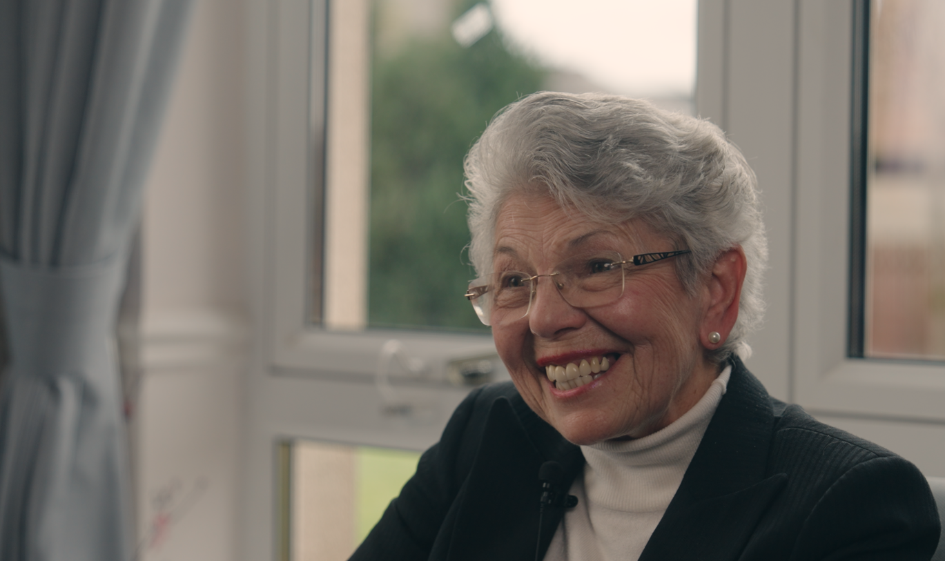Care home volunteer Liz smiles, as she sits in front of a window with a black blazer and high necked blouse 