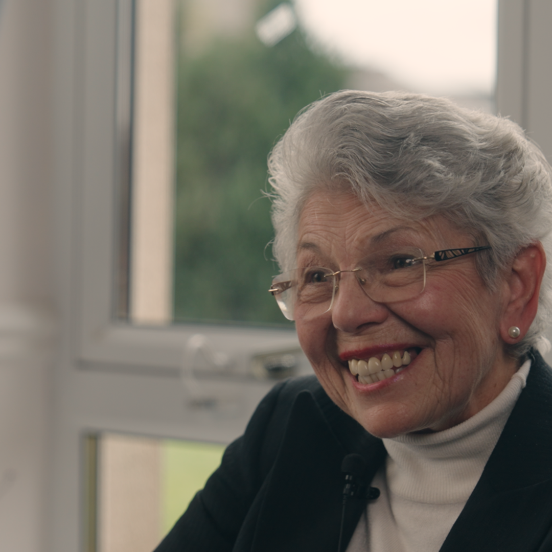 Care home volunteer Liz smiles, as she sits in front of a window with a black blazer and high necked blouse 