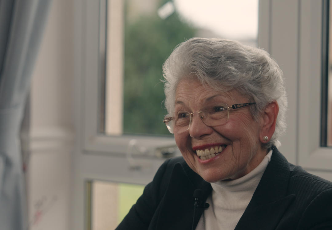 Care home volunteer Liz smiles, as she sits in front of a window with a black blazer and high necked blouse 