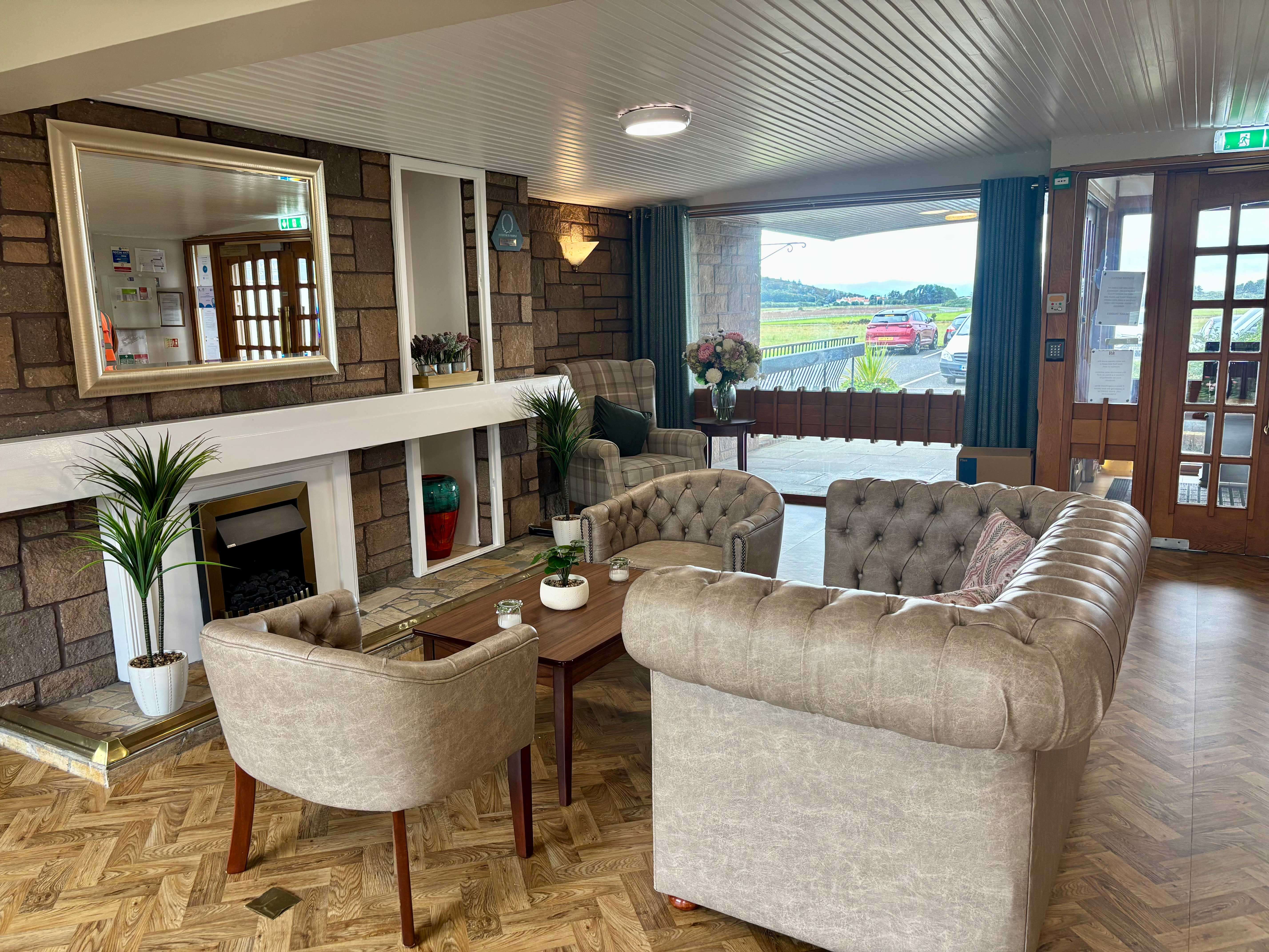A welcoming, warm-toned lounge area just inside the main entrance, featuring a large, button-tufted beige sofa and matching armchairs around a low wooden coffee table. A stone-faced fireplace is on the left, and a large window/door opens to the outside.