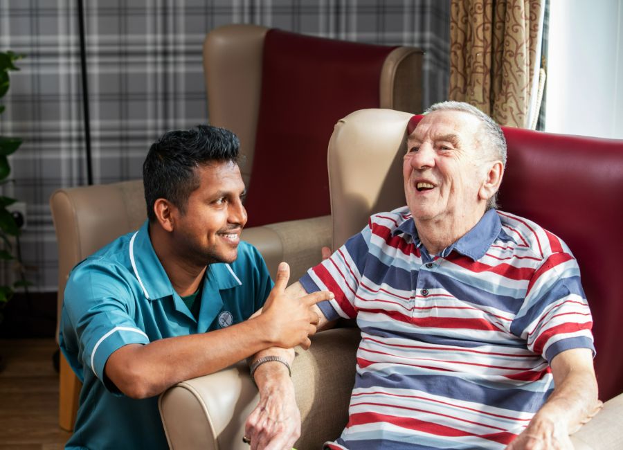 A staff member kneeling down to converse with a gentleman in a red/beige next to a large window looking out to the garden