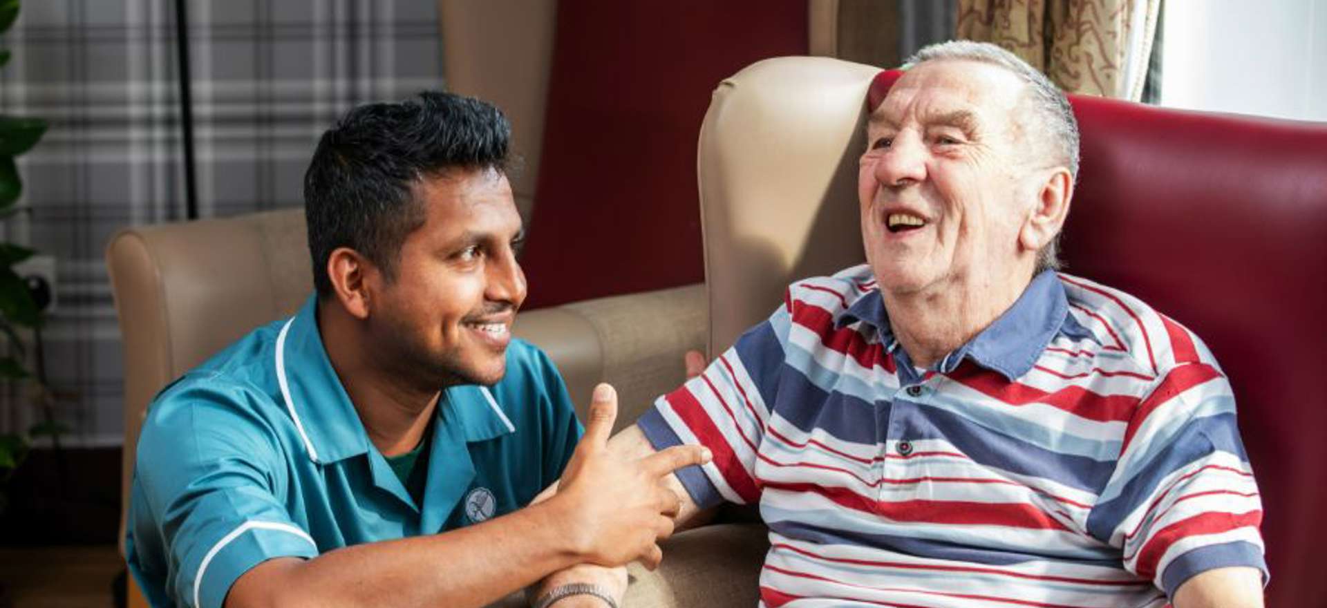 A staff member kneeling down to converse with a gentleman in a red/beige next to a large window looking out to the garden