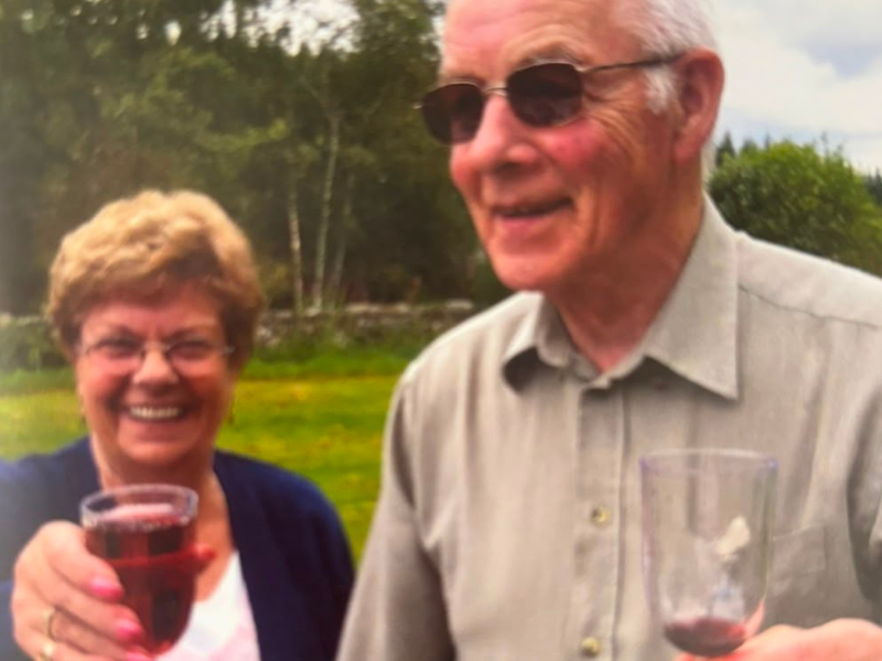 A woman stands on the left holding her glass up in a 'cheers' position. She has short, brown hair and glasses and is smiling. A man stands to her right, he is looking off to the side, smiling. He has grey hair and dark sunglasses. He is similarly holding a glass.