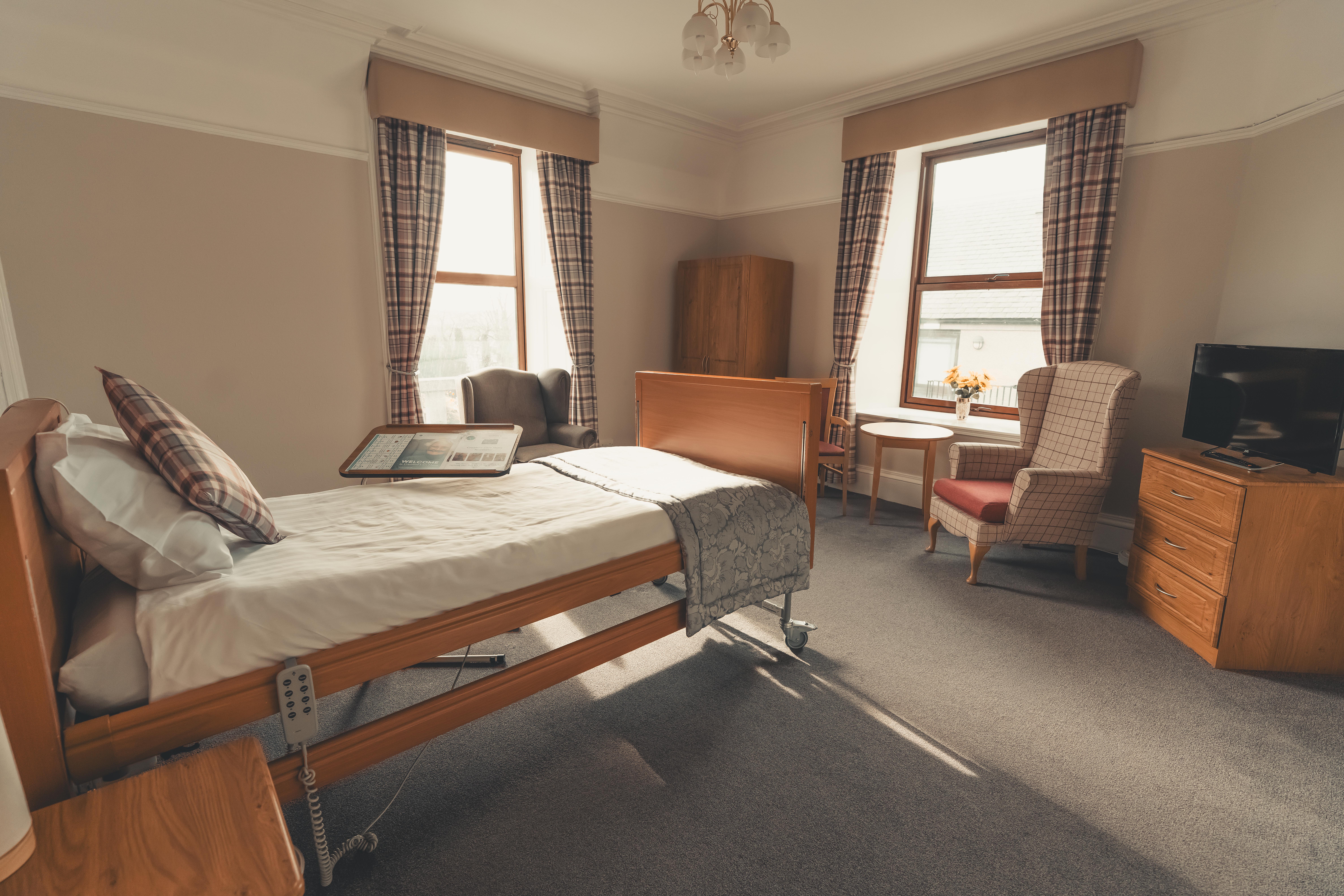 A large, bright bedroom in the care facility featuring two tall windows with plaid curtains. The room contains a wooden adjustable bed, two different styles of high-back armchairs, a chest of drawers with a TV, and a small dining table. The space is well-lit by natural light and an elegant ceiling fixture.