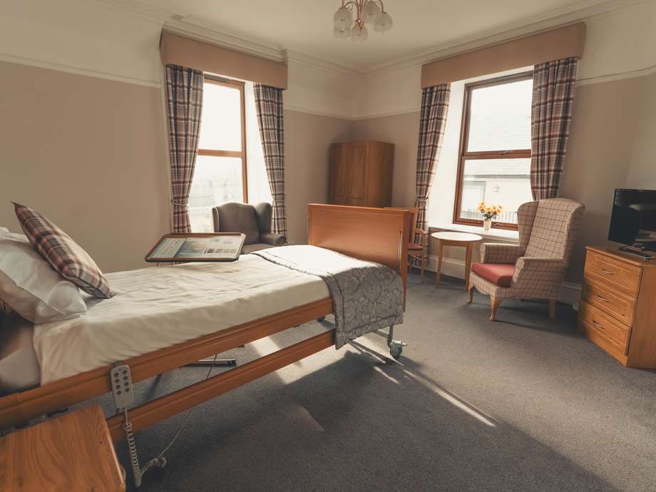 A large, bright bedroom in the care facility featuring two tall windows with plaid curtains. The room contains a wooden adjustable bed, two different styles of high-back armchairs, a chest of drawers with a TV, and a small dining table. The space is well-lit by natural light and an elegant ceiling fixture.