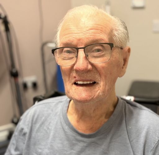 A close-up, brightly lit portrait of a man wearing rectangular, dark-rimmed glasses and a light blue t-shirt. He has white hair and a gentle smile that reveals his teeth.