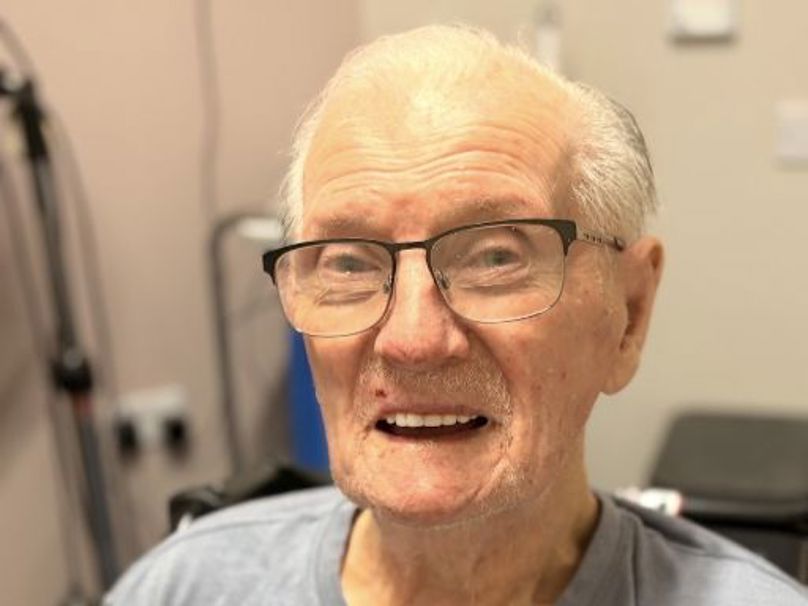 A close-up, brightly lit portrait of a man wearing rectangular, dark-rimmed glasses and a light blue t-shirt. He has white hair and a gentle smile that reveals his teeth.