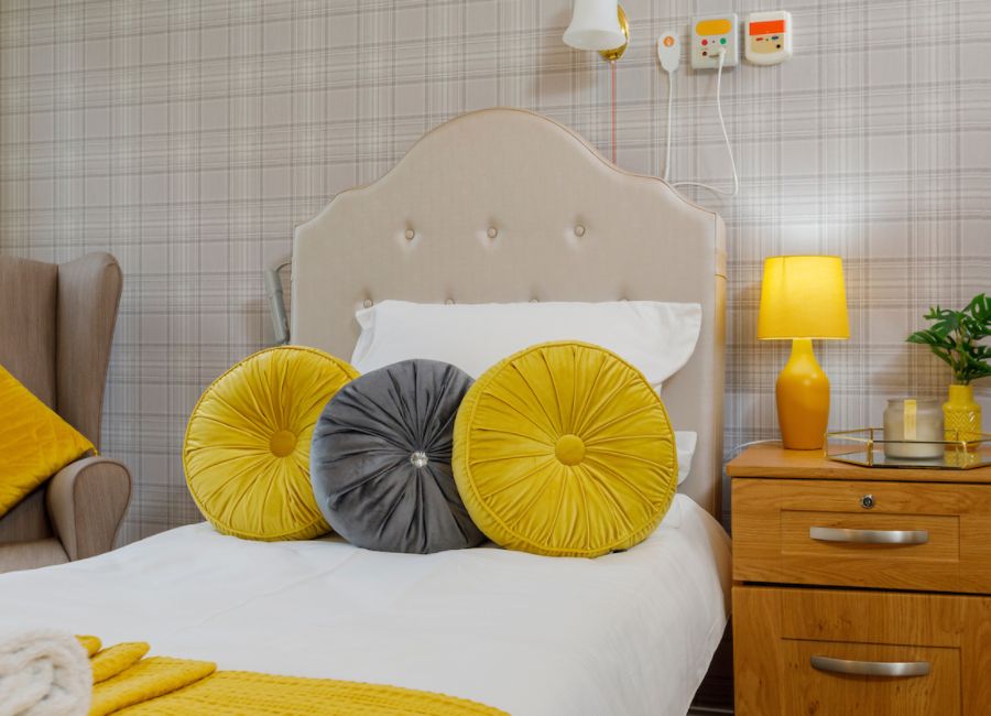 A bedroom set up from the home. Grey and beige tartan wallpaper lines the walls, with a neutral bed and chair and cosy yellow accents throughout