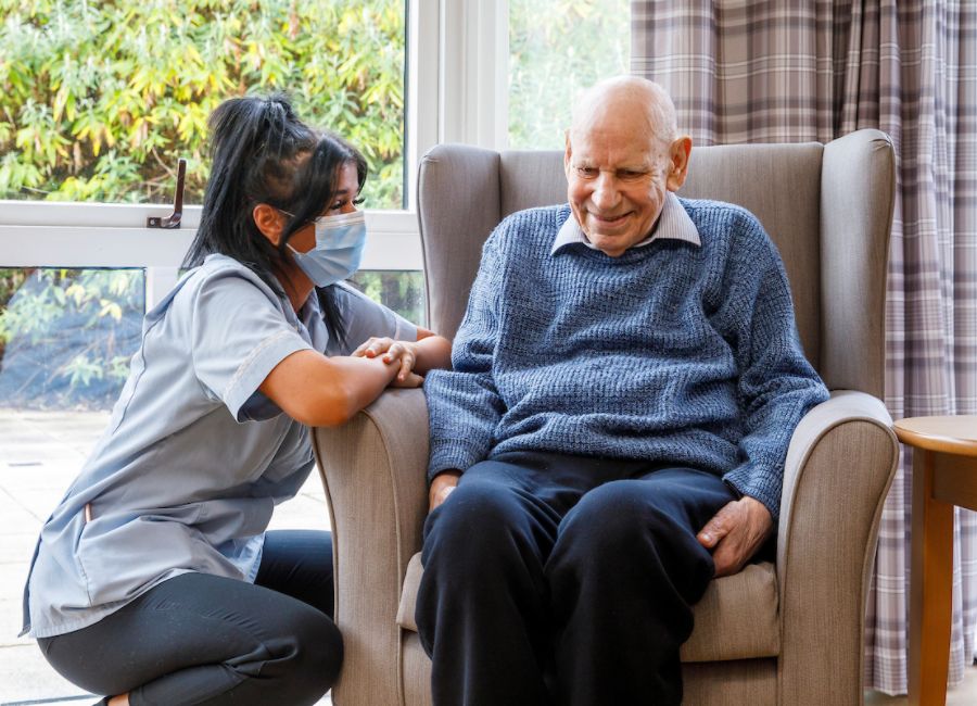A staff member kneeling down to converse with a gentleman in a grey chair next to a large window looking out to the garden