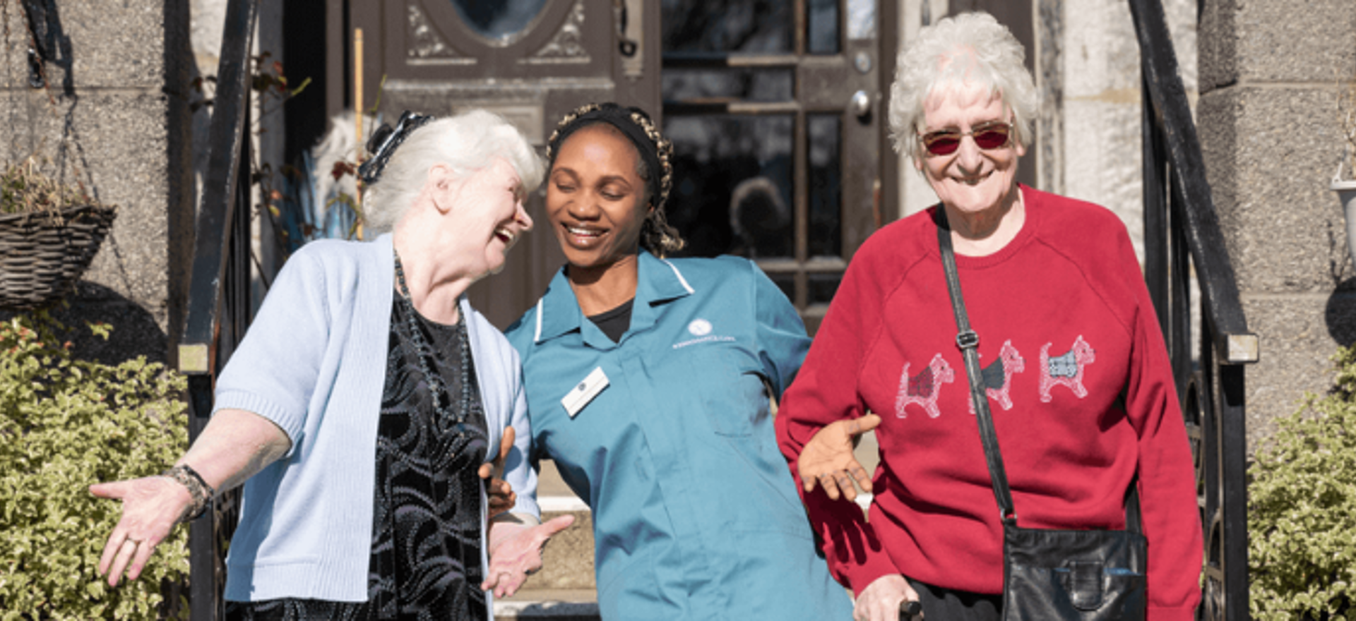 A staff member links arms with two residents, laughing along together outside the care home