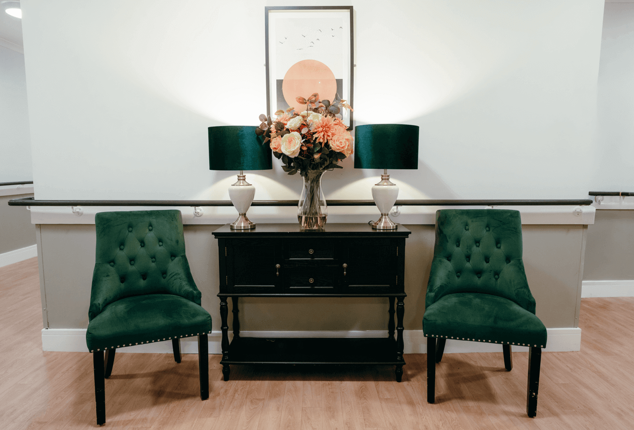 Care home hallway with two green chairs either side of a black console table. Atop the table sits two elegant lamps and a central vase filled with flowers. 