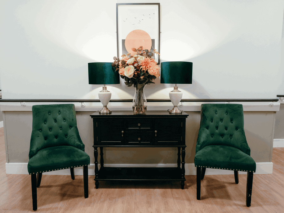 Care home hallway with two green chairs either side of a black console table. Atop the table sits two elegant lamps and a central vase filled with flowers. 
