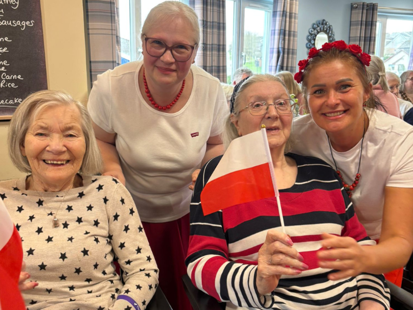 Care home residents celebrate different cultures. Two residents are holding Polish flags, with two staff members standing behind them in cultural dress.
