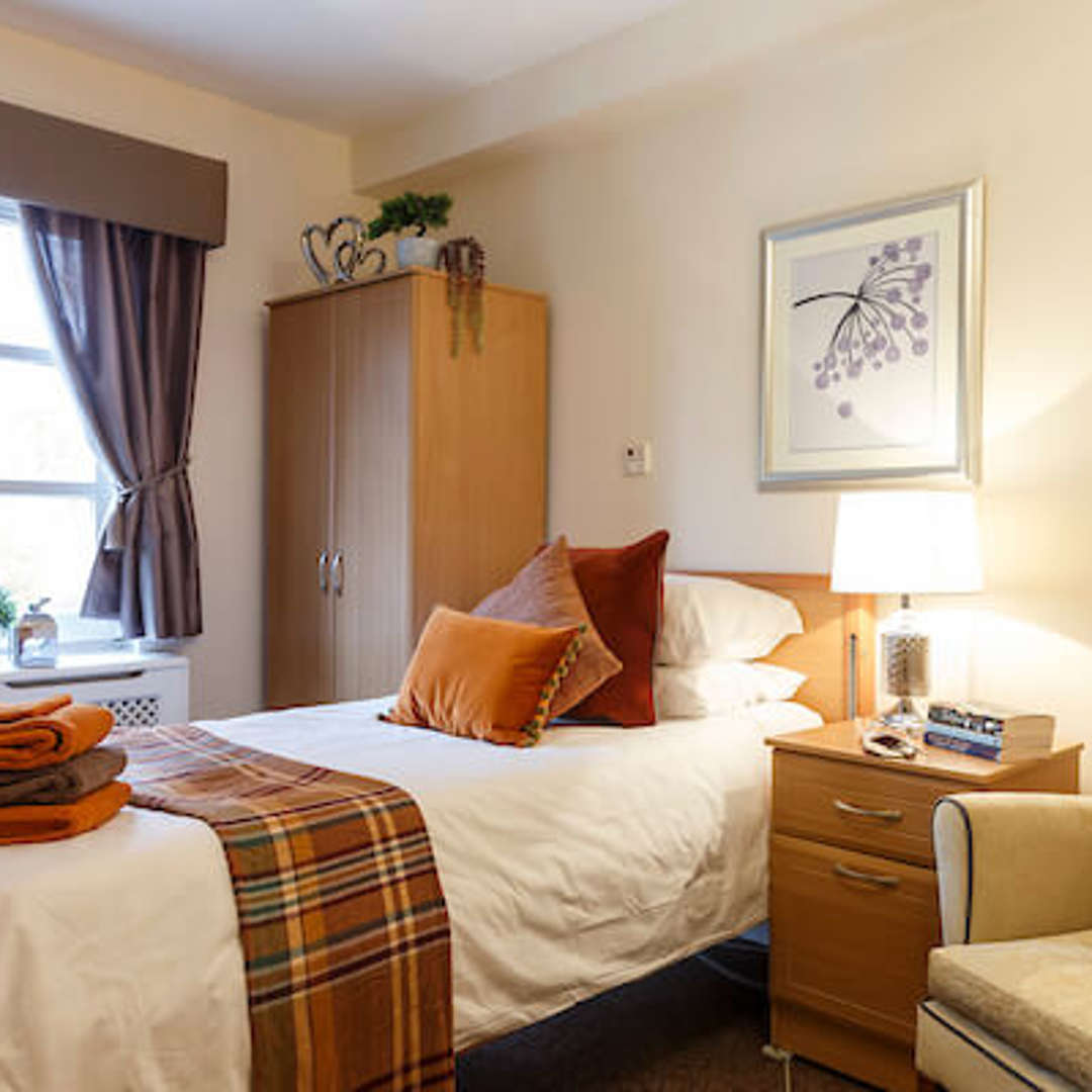 A cosy bedroom with wooden furniture. Autumn tones feature throughout, with a selection of home furnishings such as cushions, throws, and ambient lighting