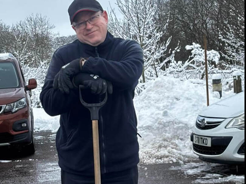 A man wearing a fleece and cap stands with a snow shovel in the care homes car park