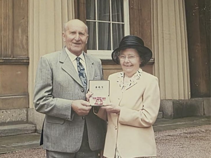 Jean Dunlop stands next to her husband as the pair hold open a presentation box displaying her MBE medal 