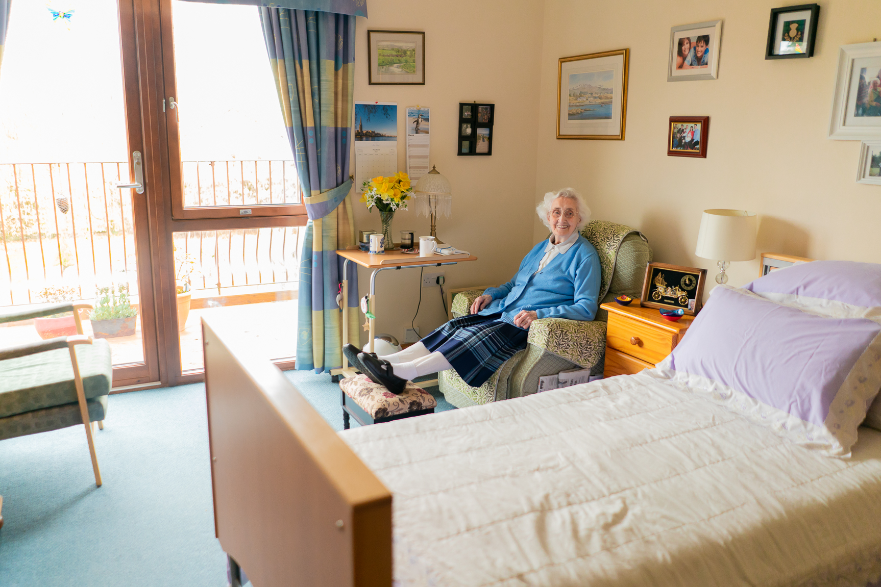 An older adult relaxes in a bedroom chair, enjoying a cup of tea and the view from the window
