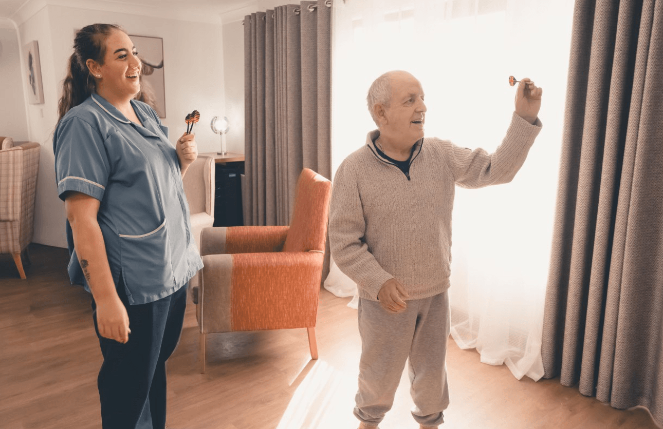 An elderly man standing in a sunlit lounge, aiming a dart toward an off-screen board. A female caregiver in a blue uniform stands beside him, holding more darts and laughing as she encourages him. The room is warmly lit with comfortable armchairs in the background