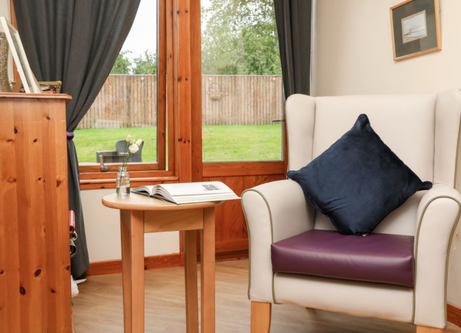 A cosy seating area in a care home. The nook has a chair with a navy blue pillow next to a small table holding an open book.